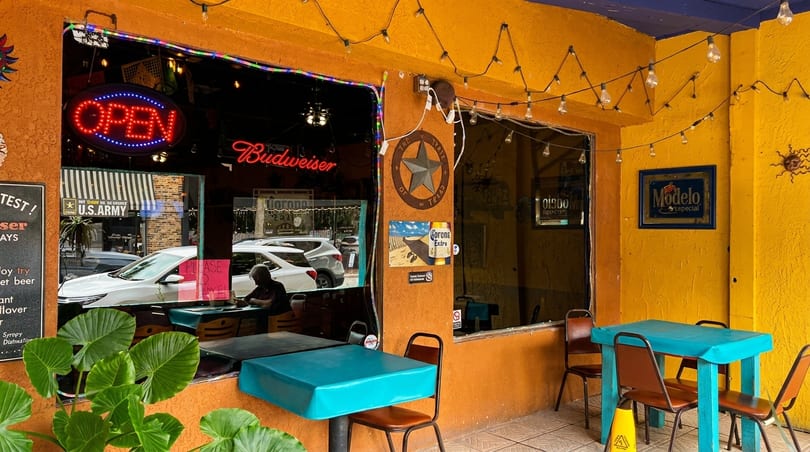 A vibrant yellow and orange restaurant exterior with an "OPEN" sign, outdoor seating, and string lights.