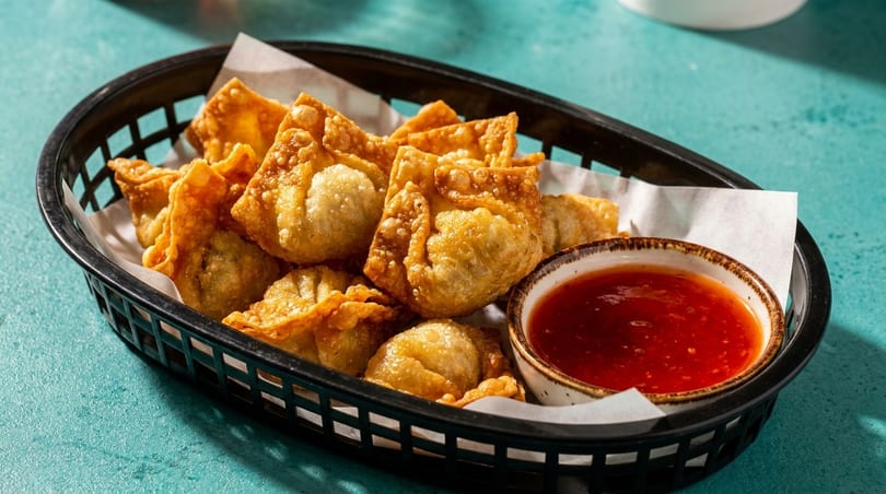 Crispy golden fried wontons in a black basket with a side of sweet chili dipping sauce.